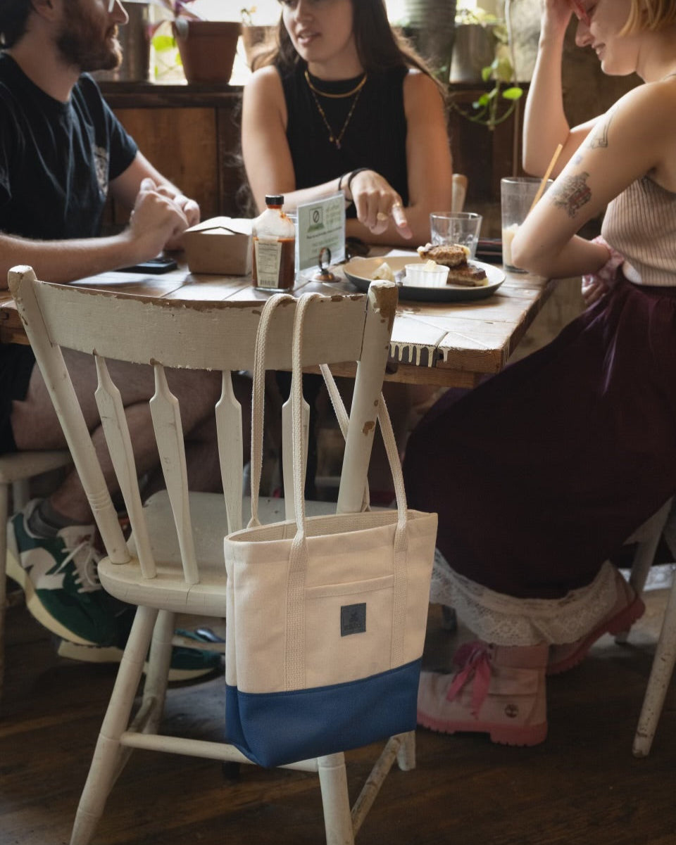 Image of models sitting around a table while the Little Everyday Tote in Blueberry + Canvas is hanging on a chair.
