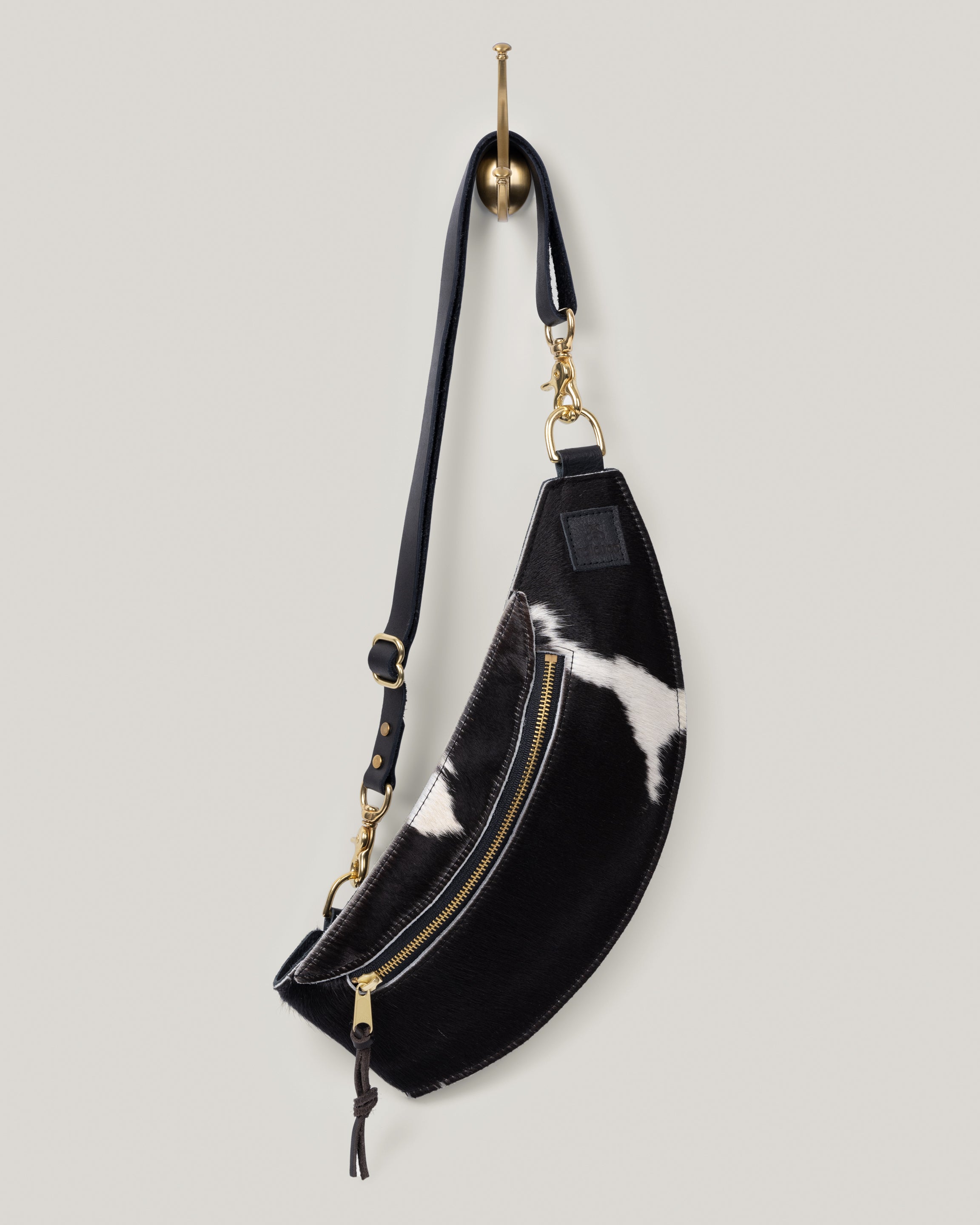 Image of the Classic Sling: Hair-on-hide in black-and-white: low profile and gently hugs your body. Just big enough to hold your phone, keys, mini wallet and chapstick. Adjustable bridle leather straps. Sturdy top of the line antique brass hardware and zipper. This Guernsey Hair-on-Hide collection gives the Classic Sling an extra level of playfulness and fun.